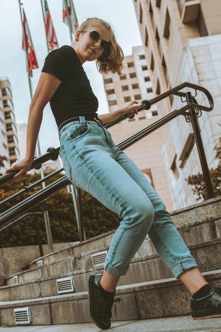 Woman posing on outdoor steps in black top, light jeans, and sunglasses in a city setting