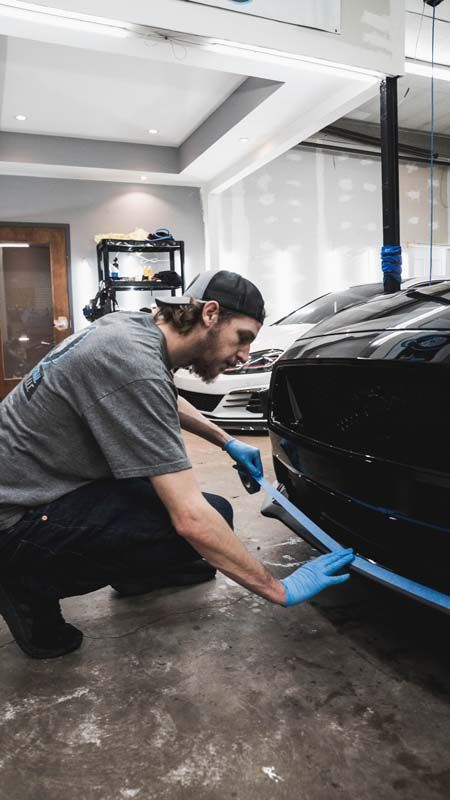 Man in a workshop applying blue tape to the front of a black car. He is wearing a hat and gloves.
