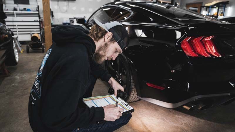 Man in a black hoodie inspects a black car's tire while holding a clipboard in a garage.