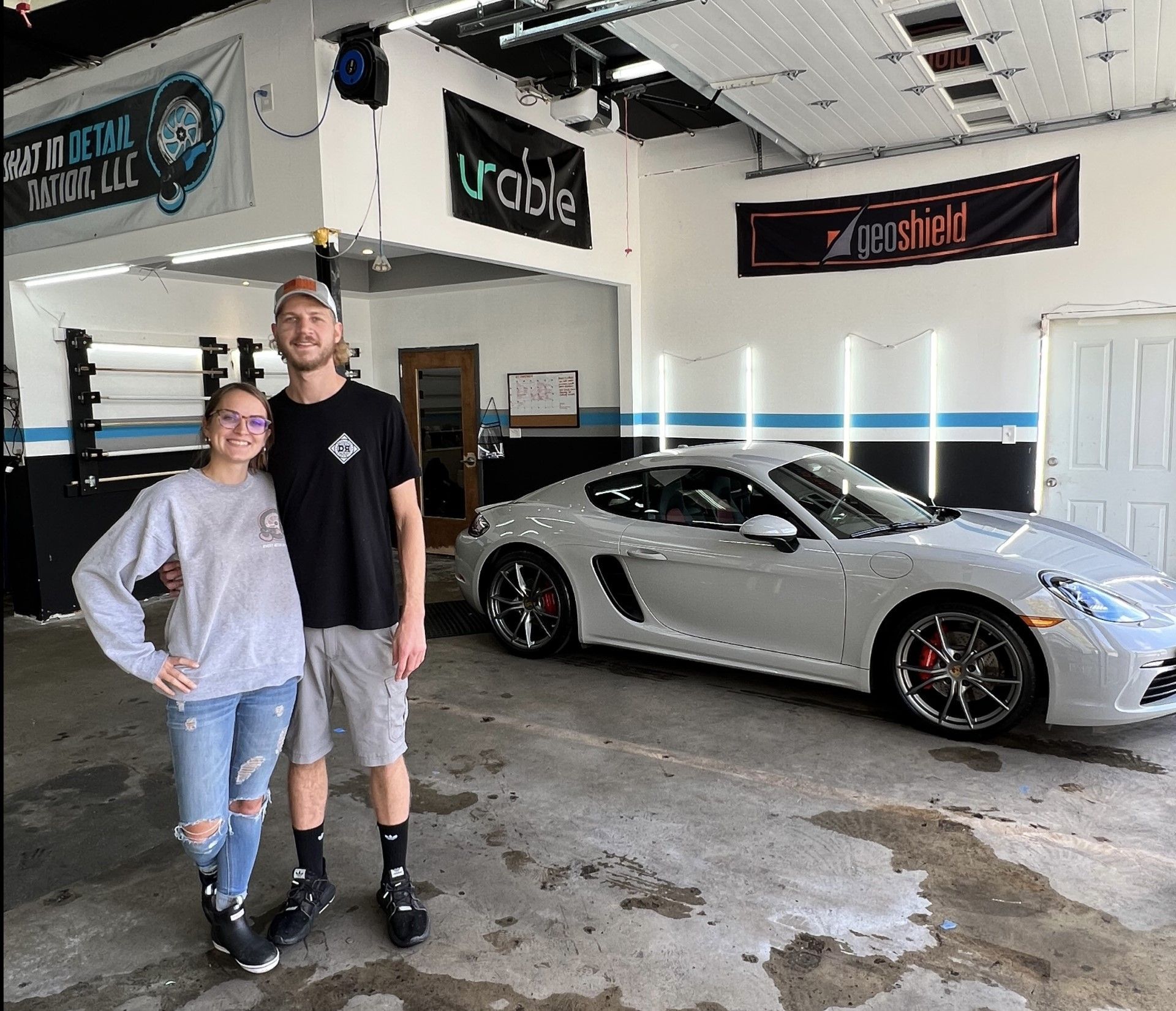 Two people stand next to a silver Porsche inside a garage.