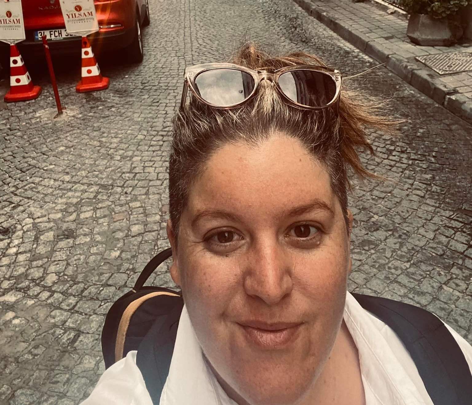 Woman with sunglasses on head takes a selfie on a cobblestone street, backpack visible.