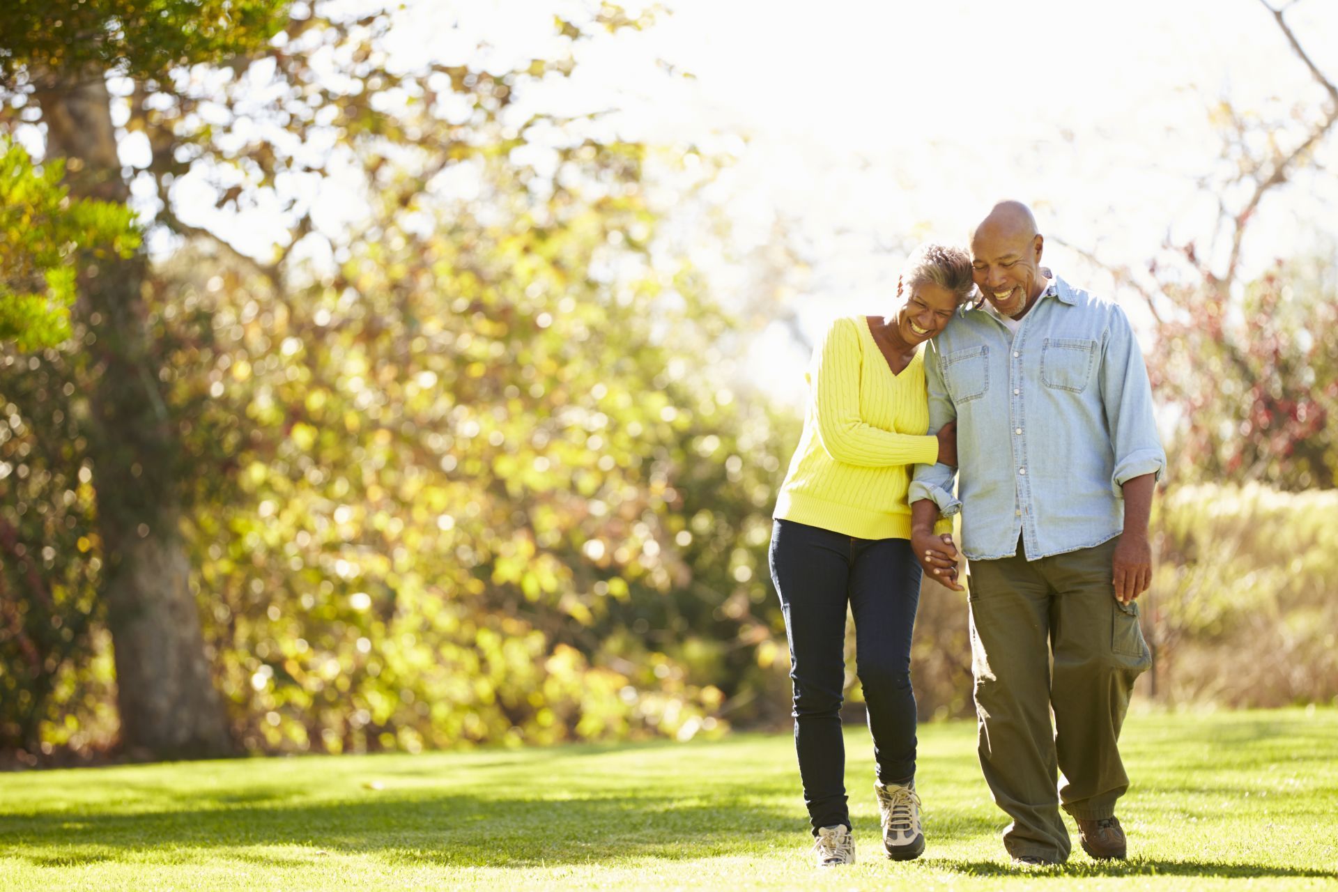 Two people walking arm in arm in a sunny park, with trees and grass in the background