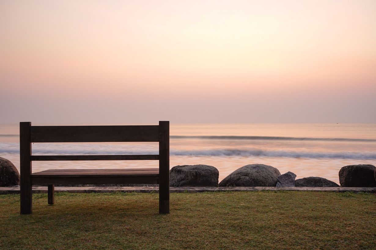 Wooden bench facing a calm ocean at sunset, with rocks and a pastel sky in the background
