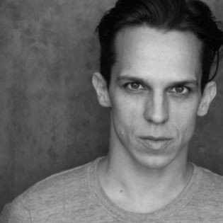 A man in a grey shirt is looking at the camera in a black and white photo.