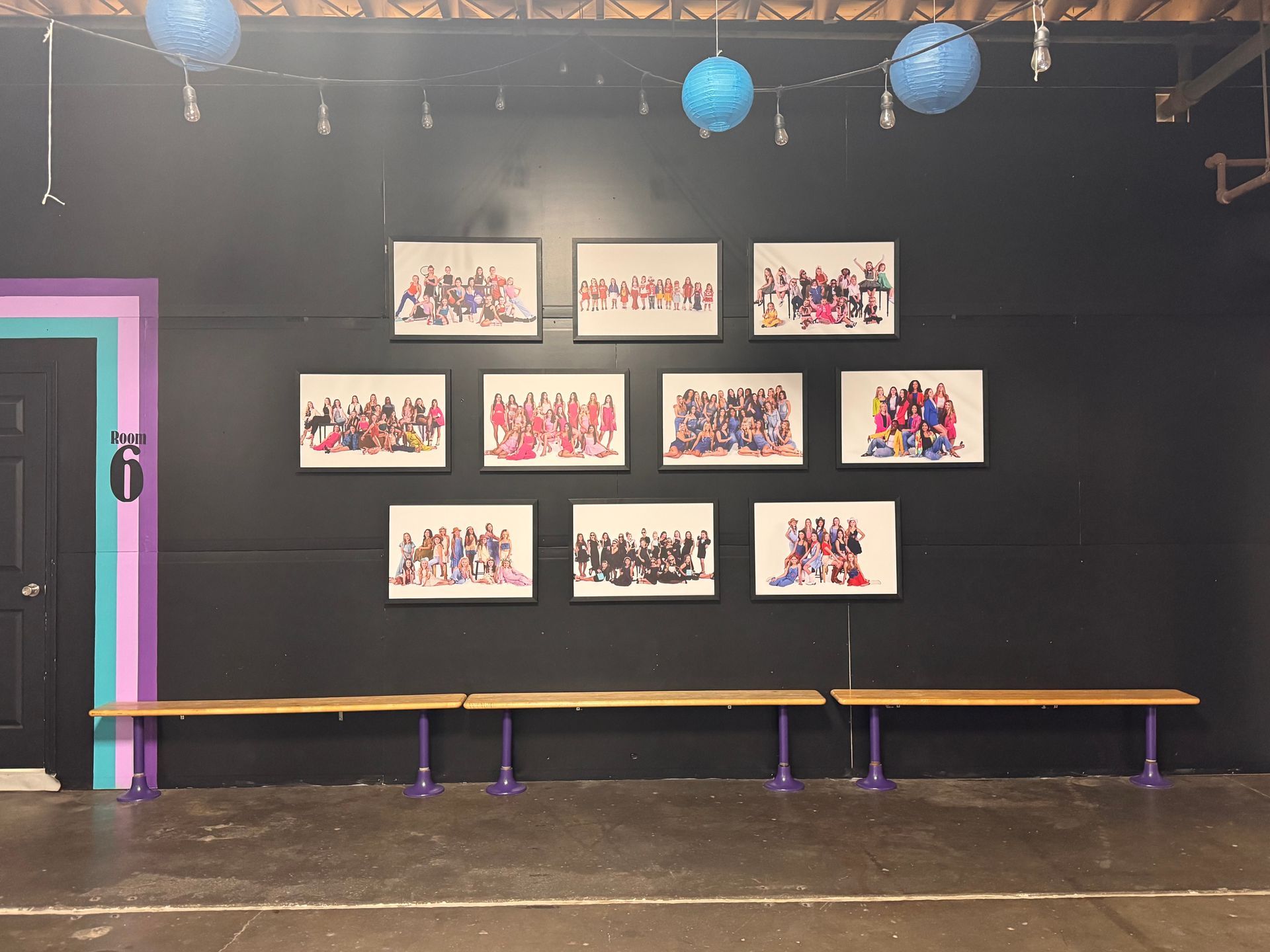 Eight framed group photographs hang on a dark wall above a long wooden bench in a room with blue paper lanterns.