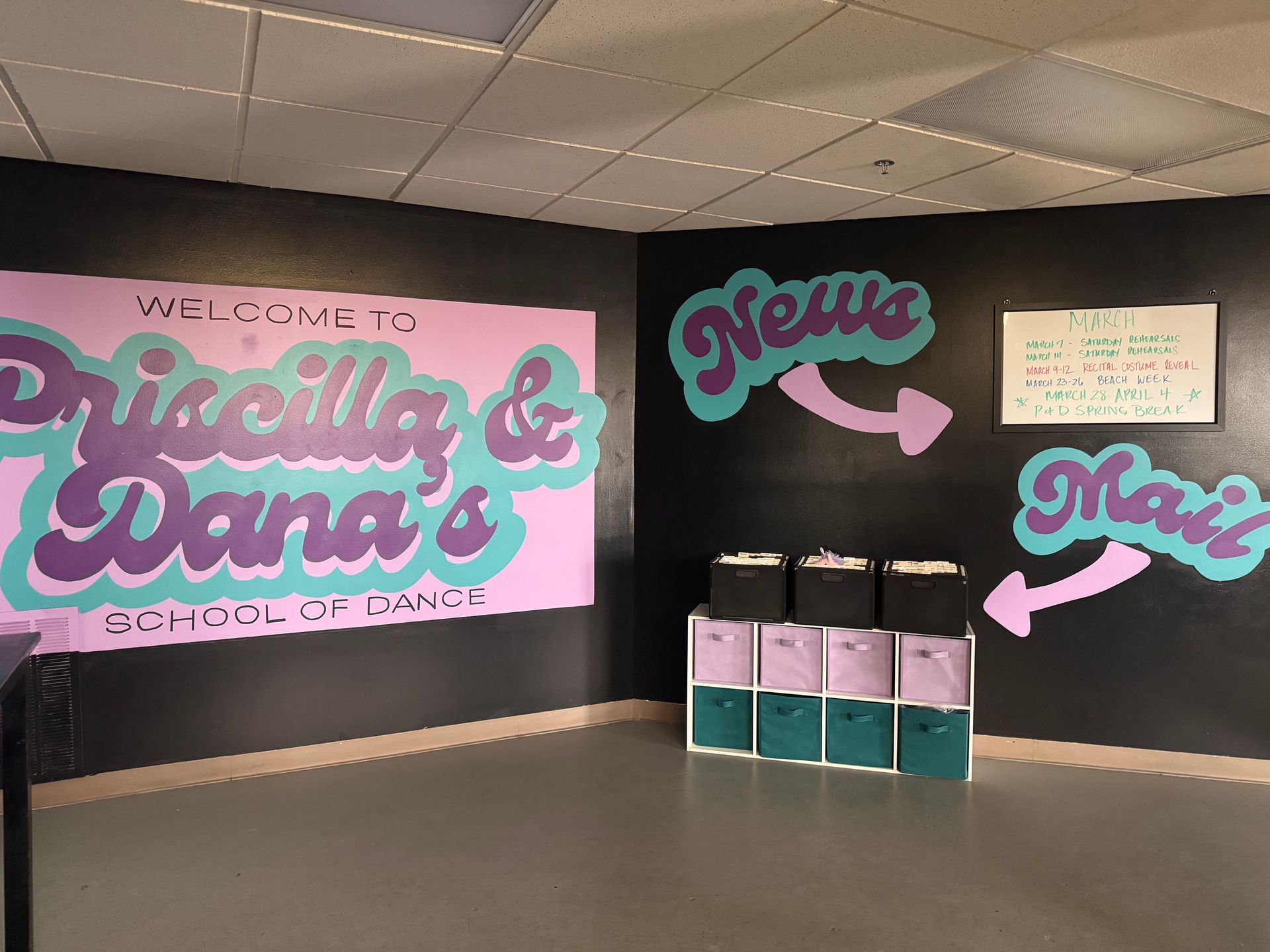 Interior of Priscilla & Dana's School of Dance with a sign, news bulletin board, and cubby storage bins on a black wall.