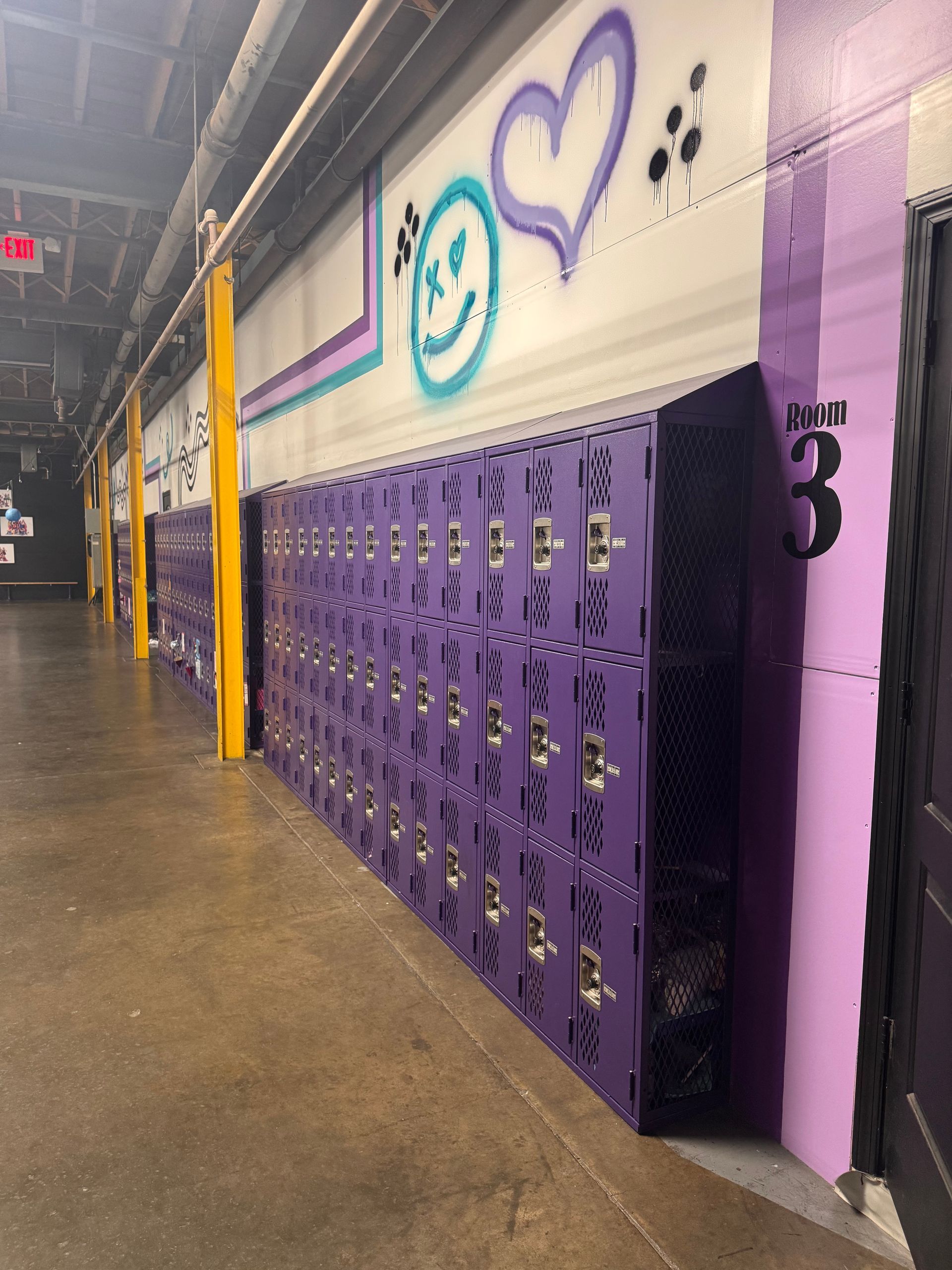 A hallway with rows of purple lockers lined against a white wall featuring graffiti art of a smiley face and a heart.