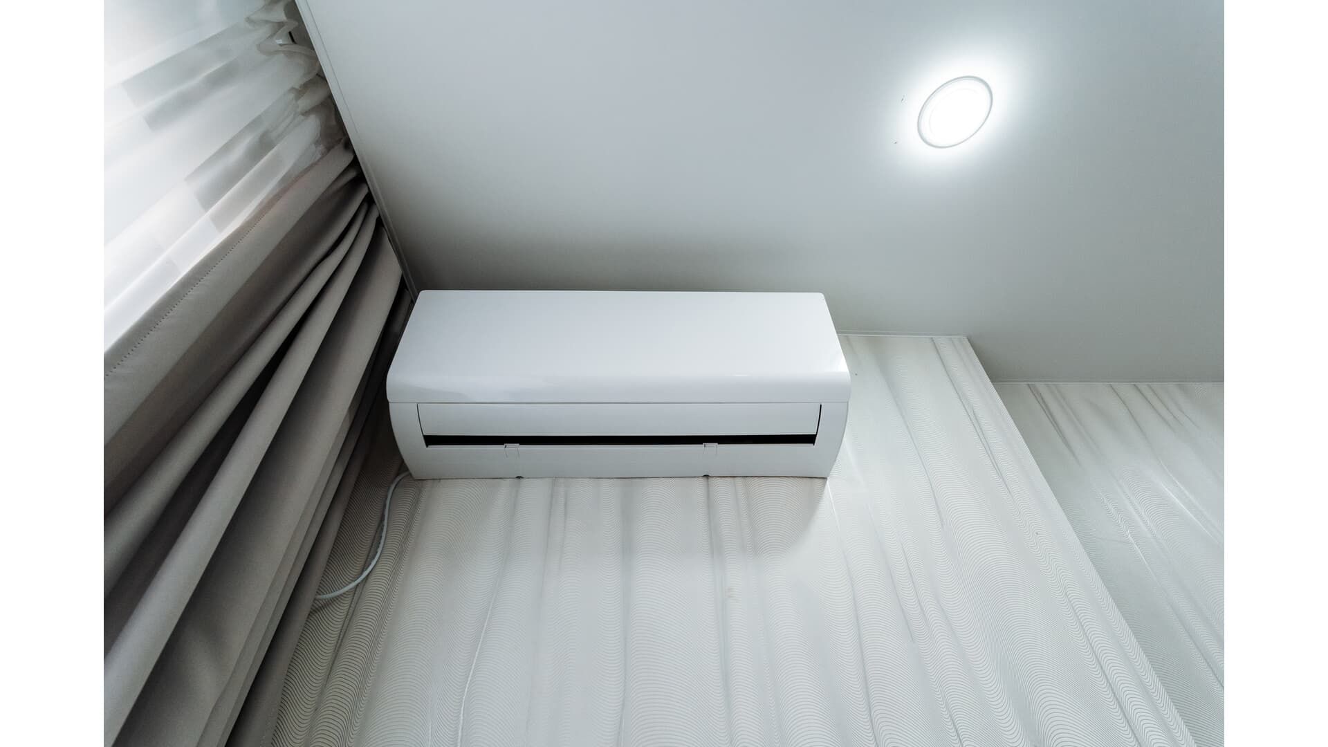 A white wall-mounted air conditioner unit installed near a ceiling and curtains.