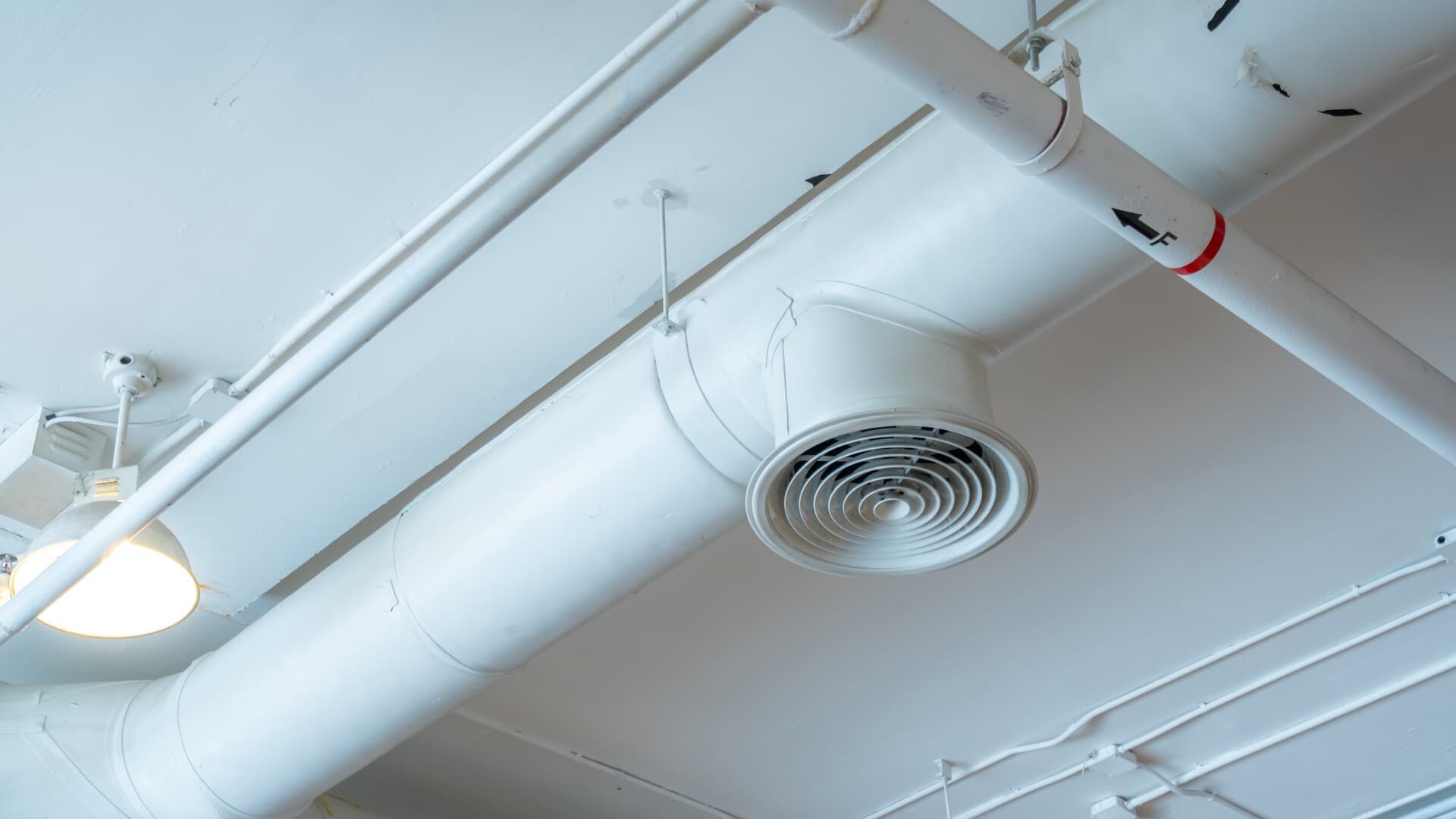 A circular white HVAC ceiling vent attached to a duct pipe and a light fixture against a white industrial-style ceiling.