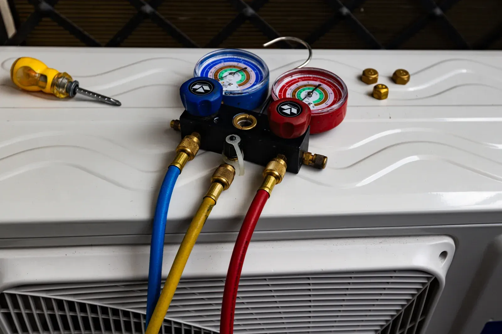 Manifold pressure gauges with blue, yellow, and red hoses connected to a white air conditioning unit with a screwdriver.
