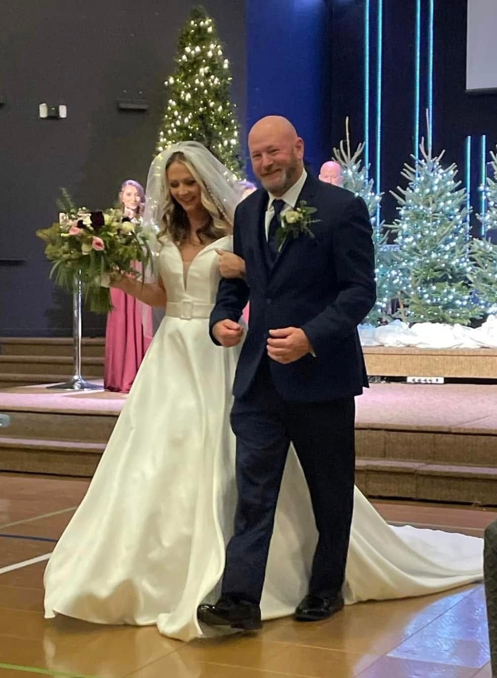 Bride and man in navy suit walking down church aisle with Christmas trees behind them