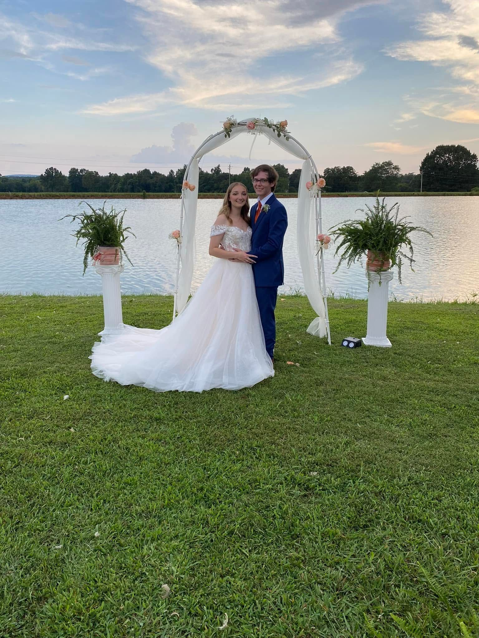 Bride and groom under a floral arch by a lakeside at sunset, standing on a grassy lawn
