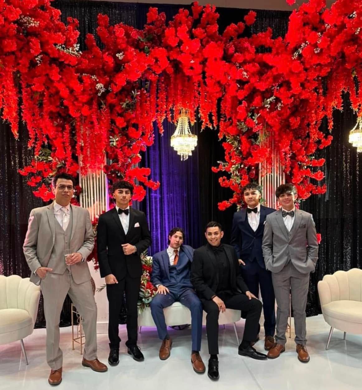 Five men posing in suits under red floral decor and chandeliers on a stage-like set