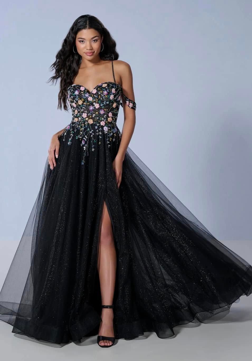 Woman in a black floral ball gown with a sheer flowing skirt, standing against a gray studio background.