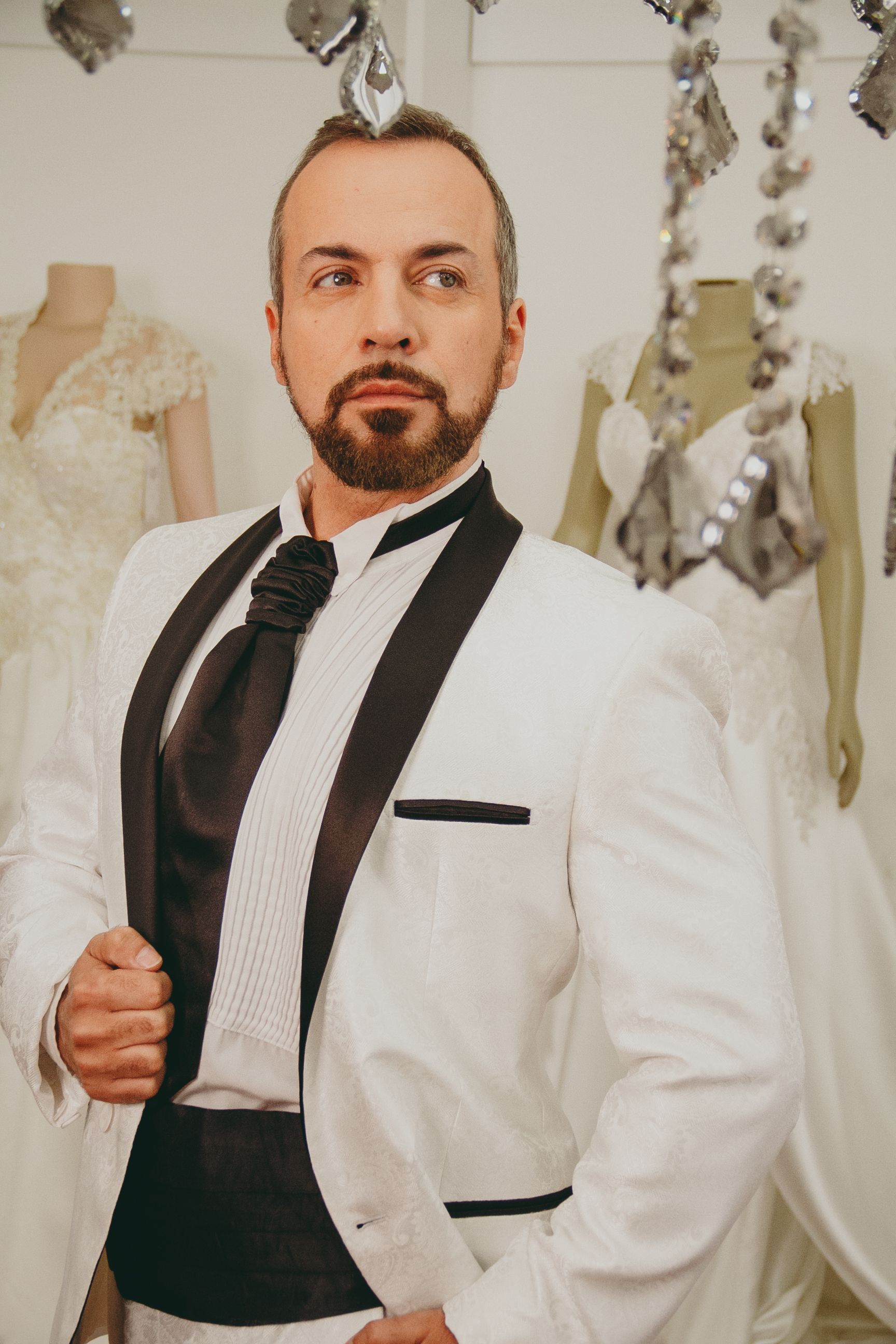 Person in a white tuxedo jacket and black tie standing indoors, holding the lapel, with decorative hanging ornaments behind.