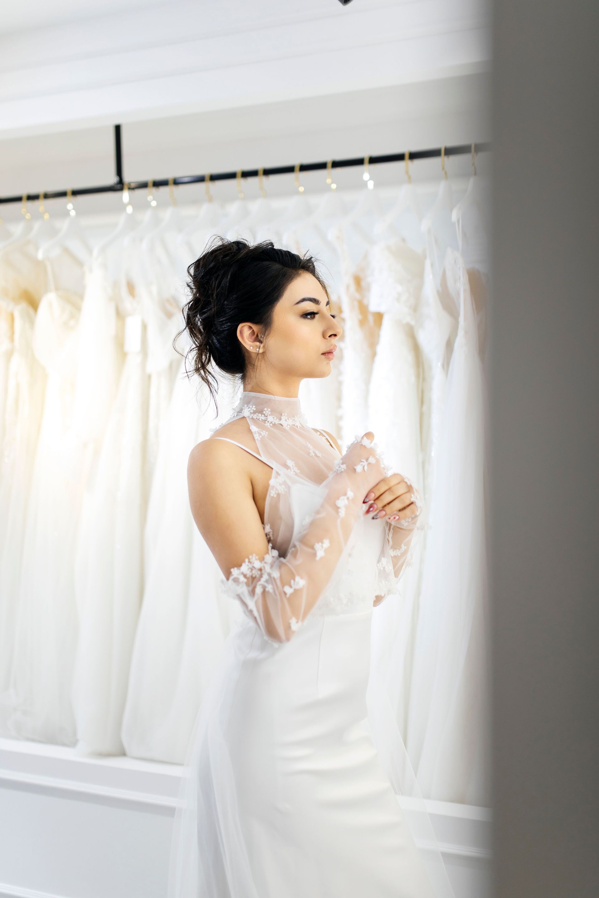 Woman in a white dress standing before a rack of wedding gowns, holding a sheer lace veil.