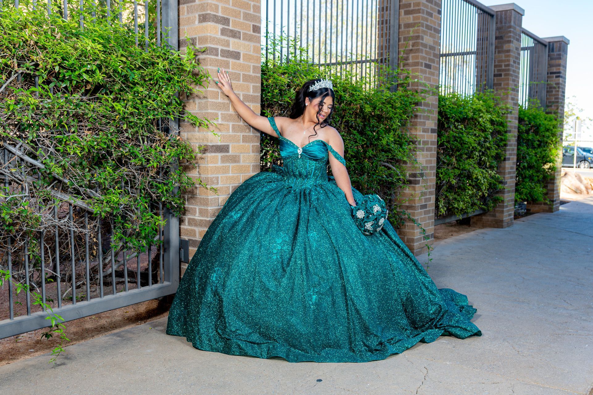 Person in a teal ballgown posing beside a brick wall and hedge-lined sidewalk