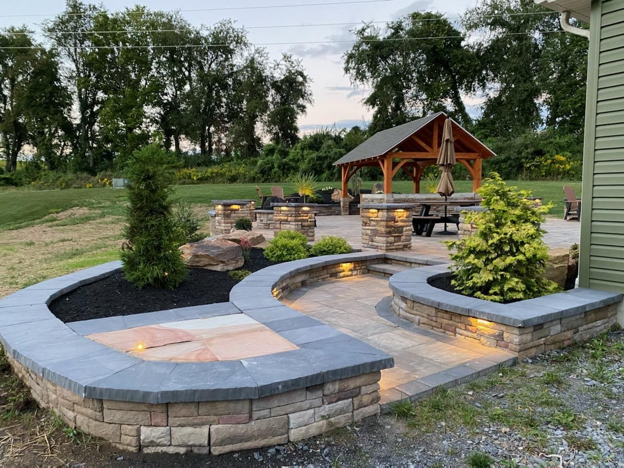 Curved stone seating around a fire pit in a landscaped backyard with a wooden gazebo and trees beyond