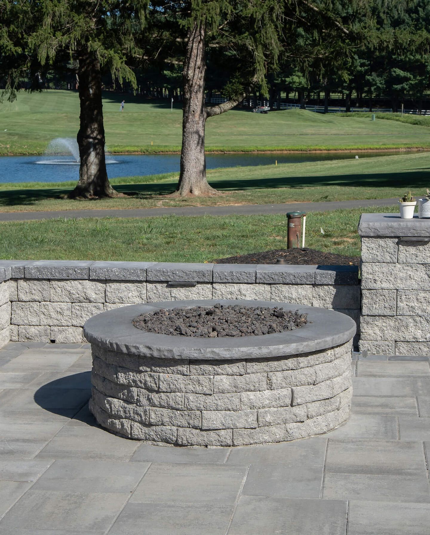 Stone fire pit in a paved park area, with a low stone wall and grassy field beyond.