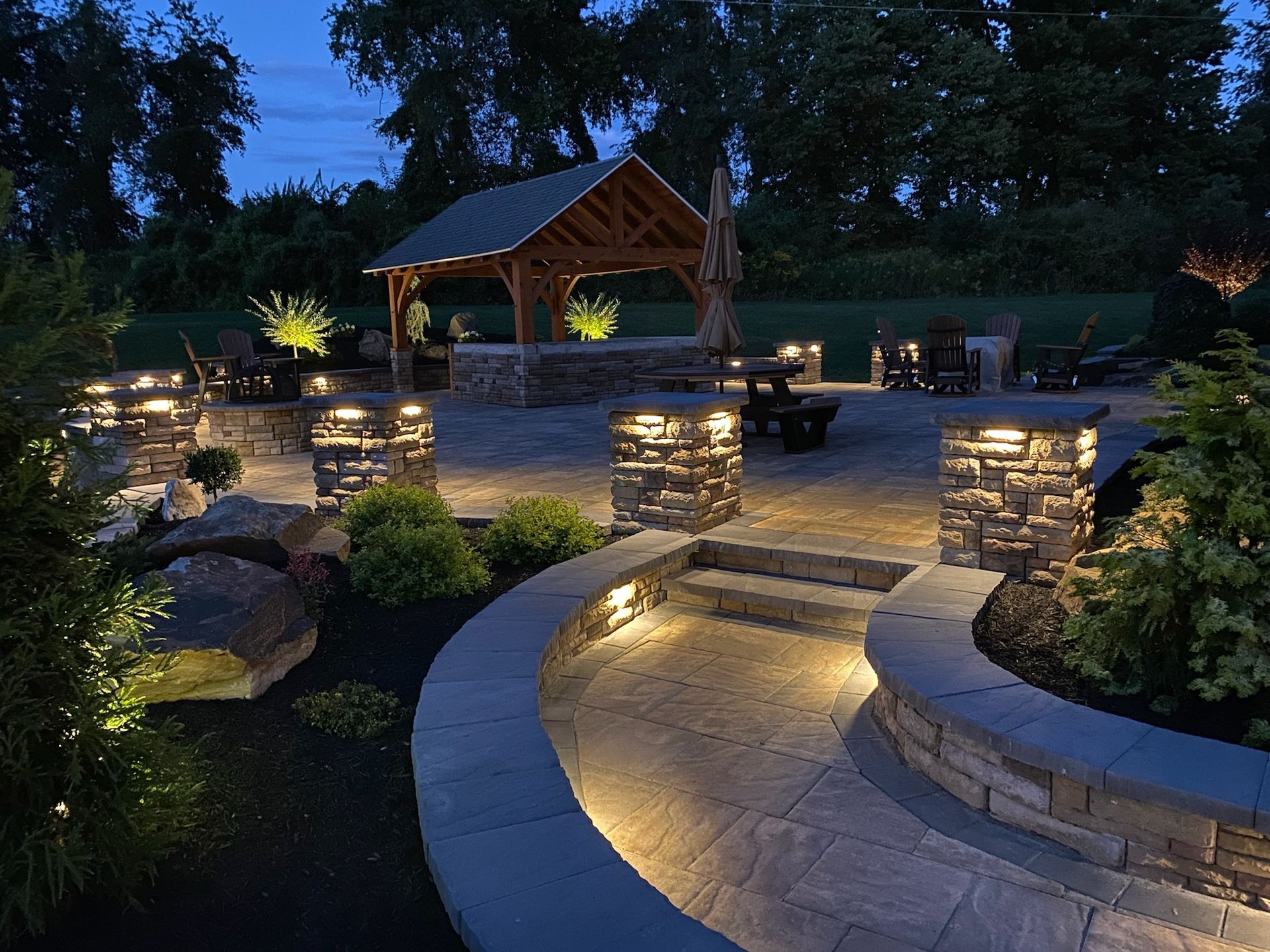 Illuminated stone patio with curved steps, landscaped beds, and a wooden gazebo at dusk