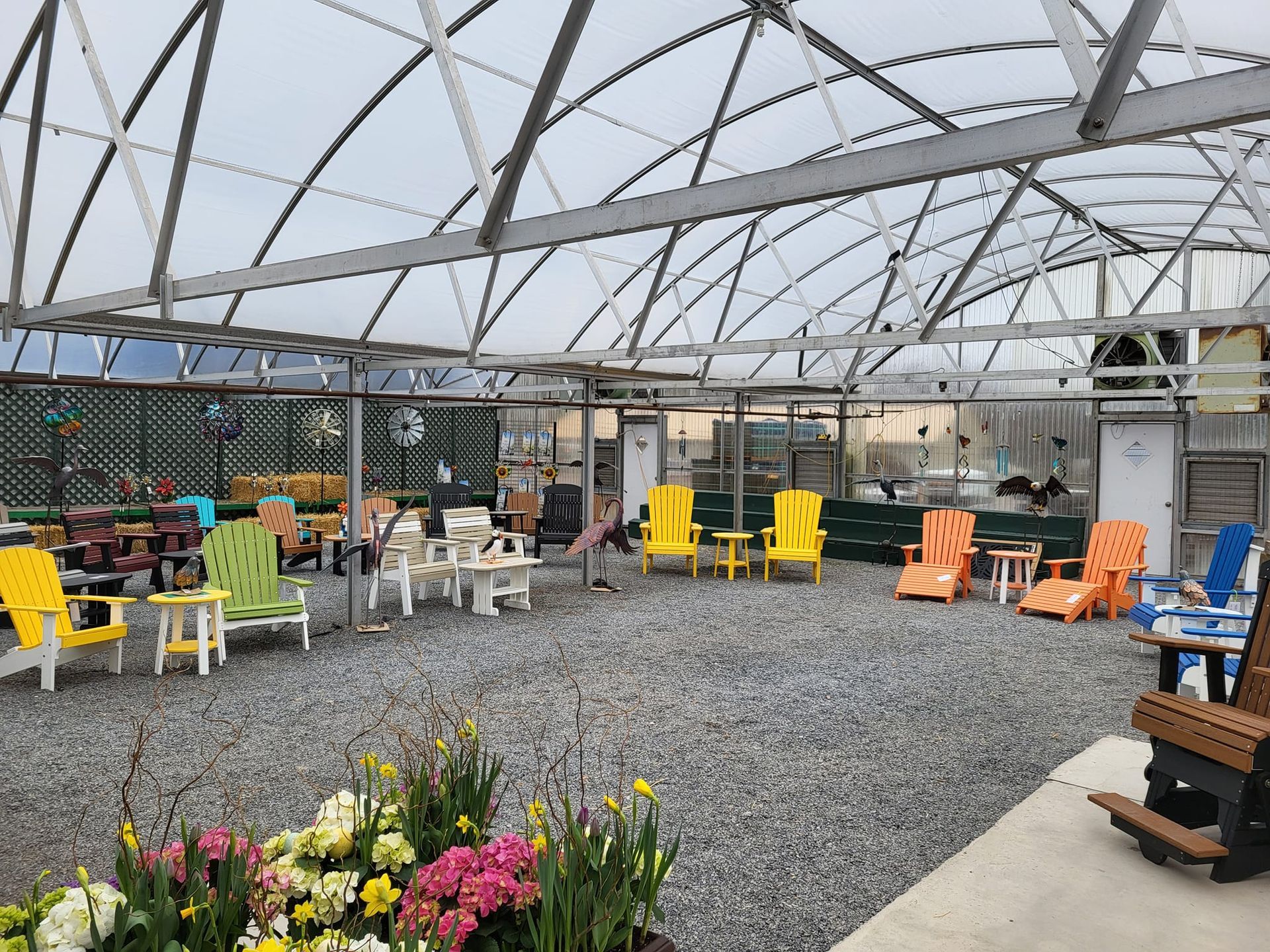 Colorful chairs and tables inside a glass-roofed greenhouse with gravel flooring and potted flowers.