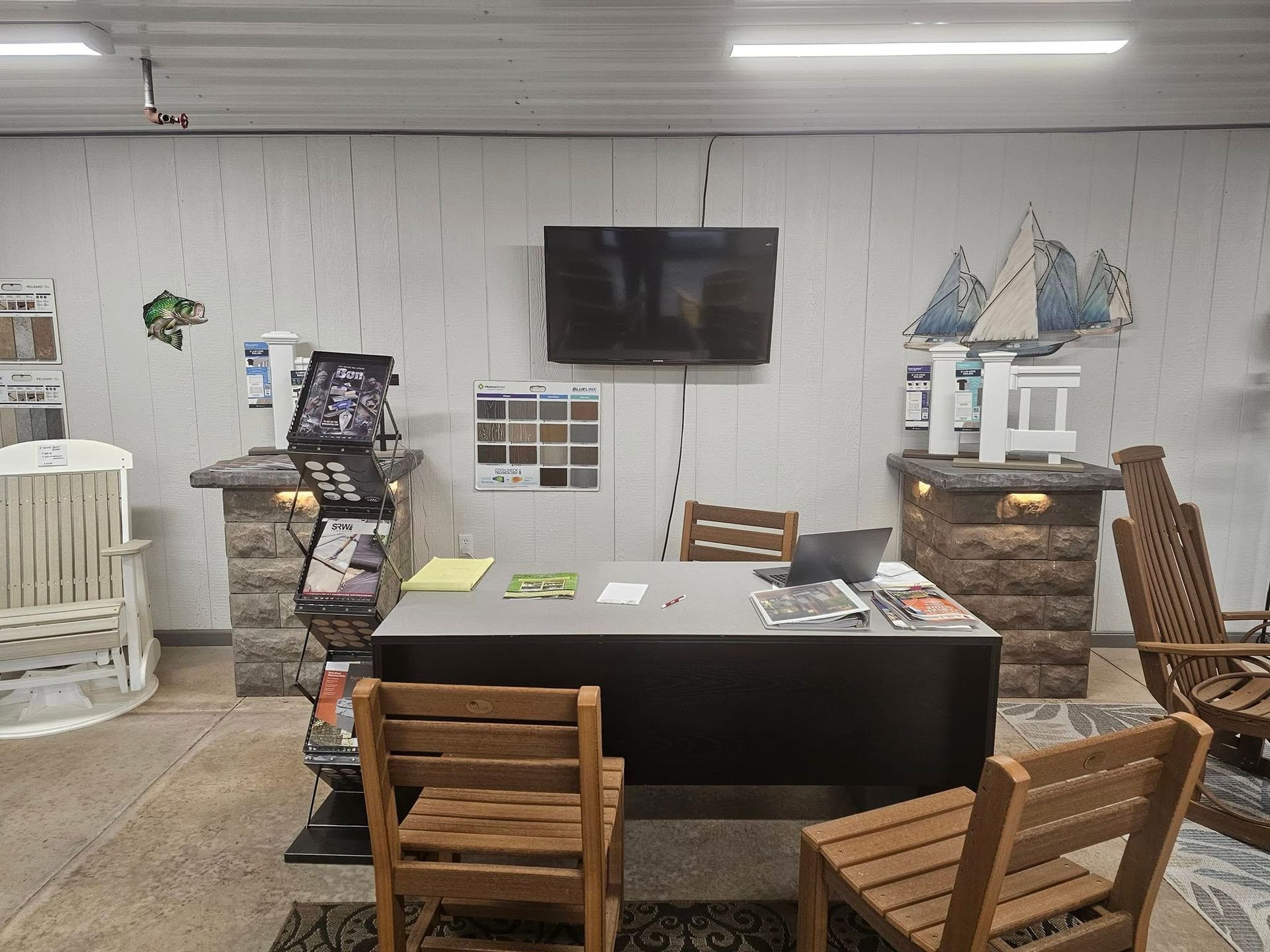 Small showroom with a black desk, wooden chairs, wall-mounted TV, and shelves on a gray wall.