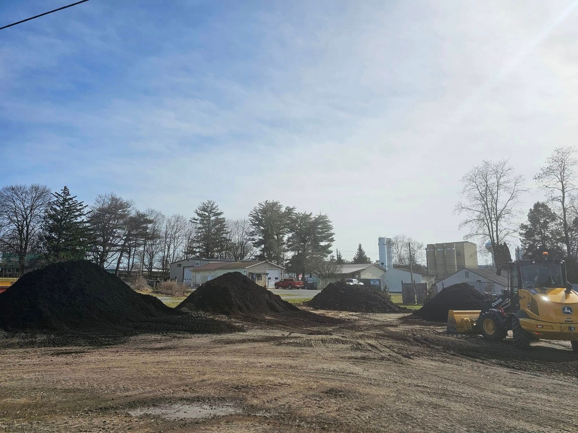 Construction site with dirt piles, trees, and yellow machinery under a cloudy sky