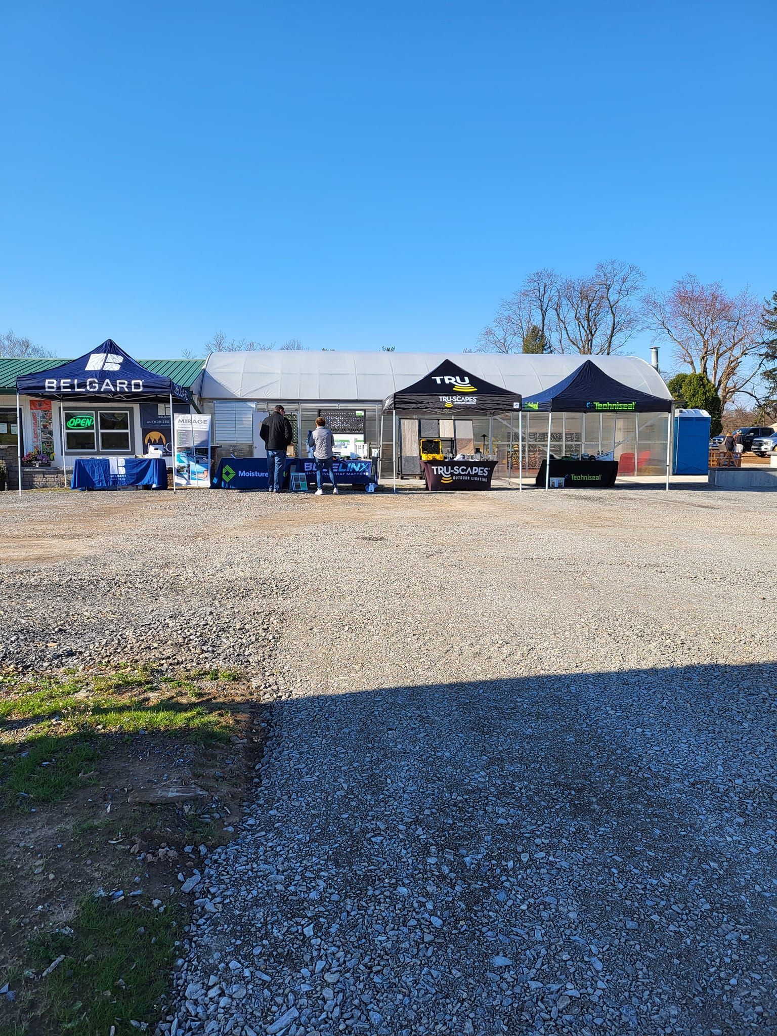Outdoor market stalls with blue canopies on a gravel lot under a clear blue sky.