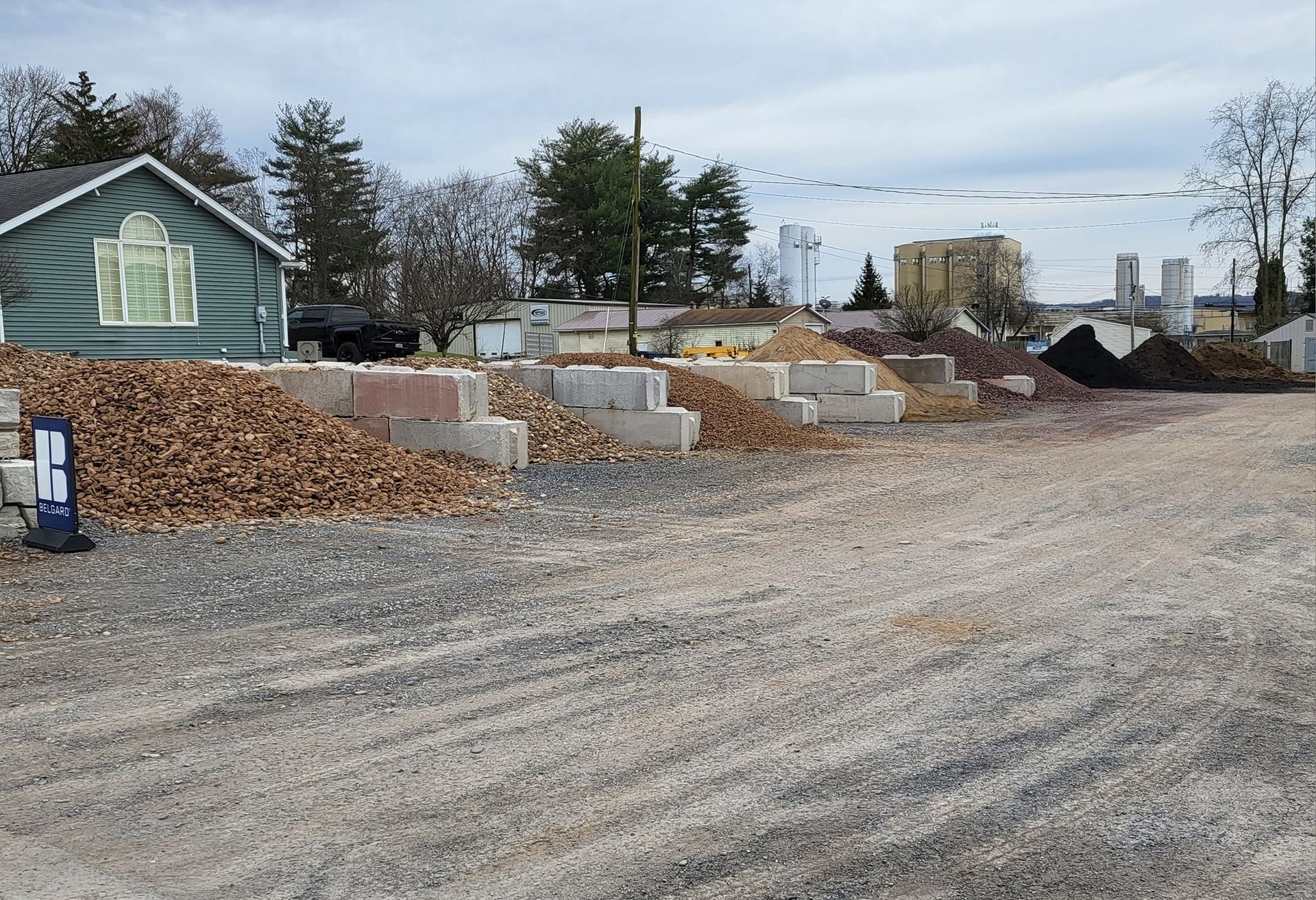 Outdoor gravel lot with stacked concrete blocks, wood chips, and a green house under cloudy skies