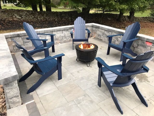 Four blue Adirondack chairs around a small fire pit in a stone patio seating area.
