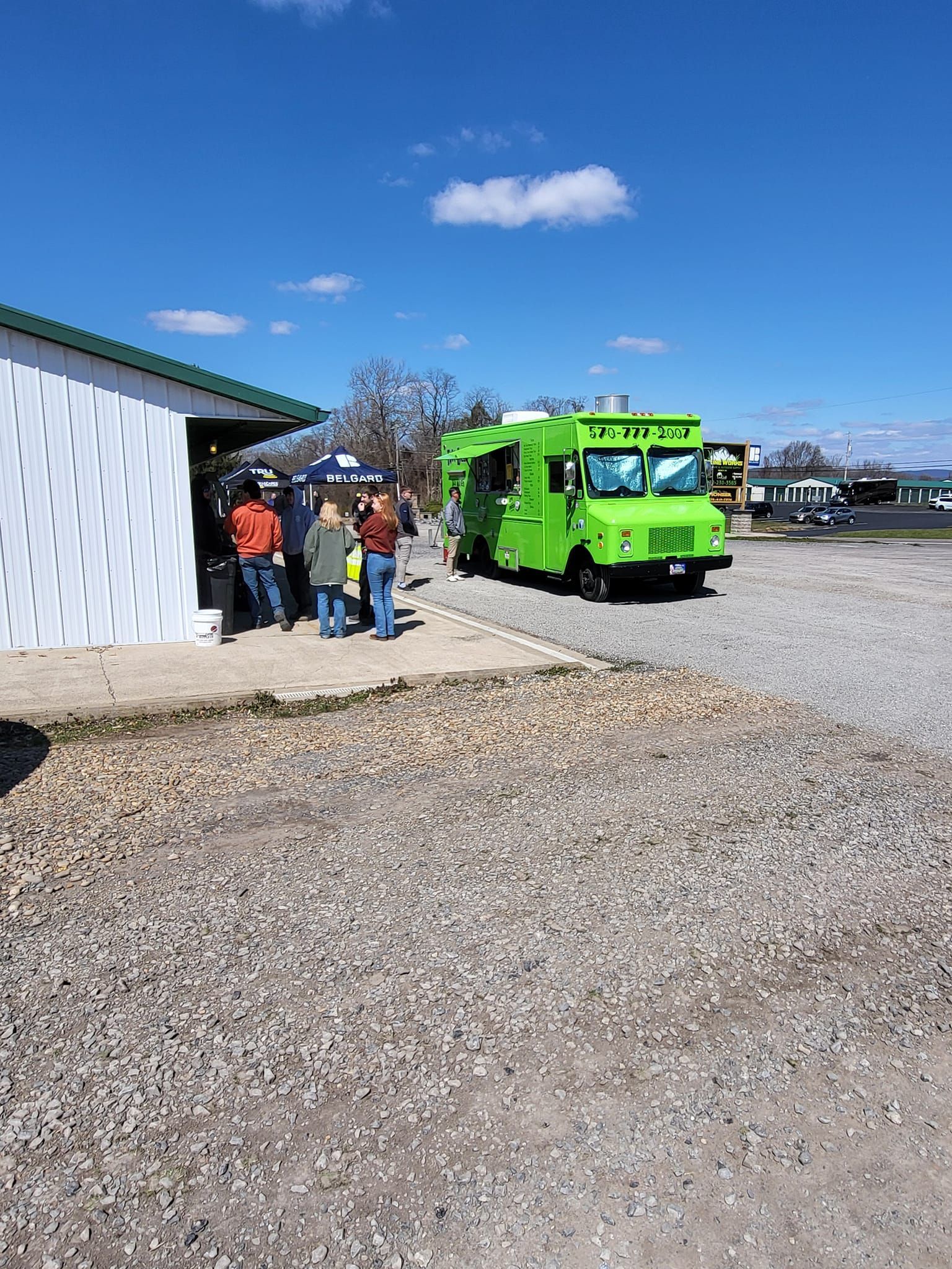People standing by a bright green truck beside a white building on a gravel lot under a blue sky