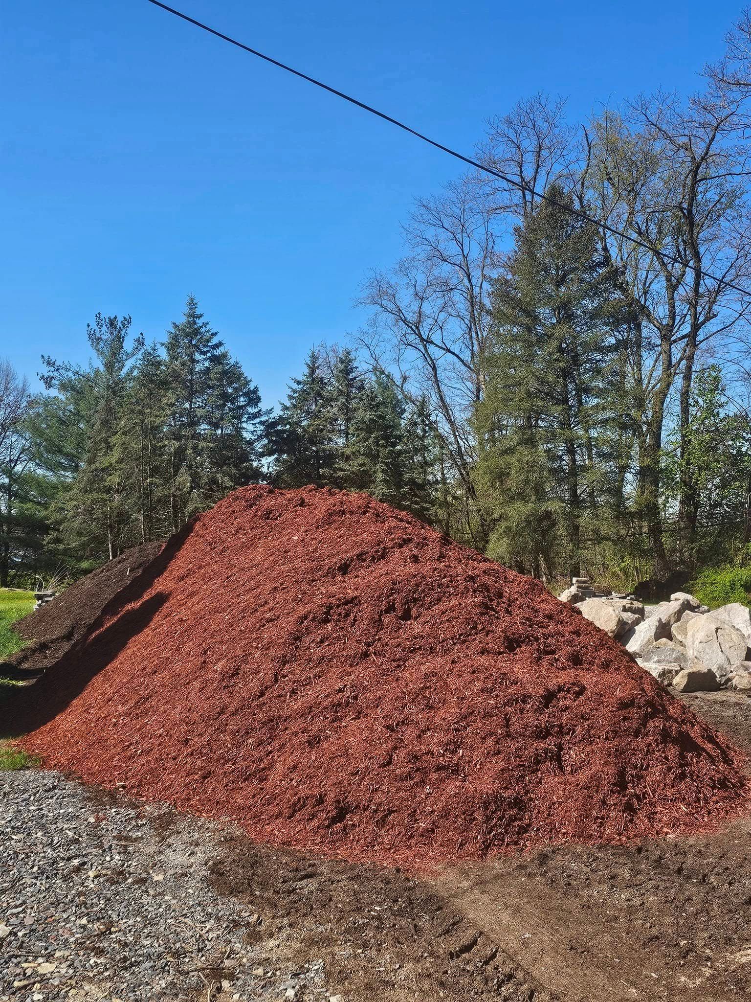 Large pile of red mulch outdoors beside gravel and trees under a blue sky