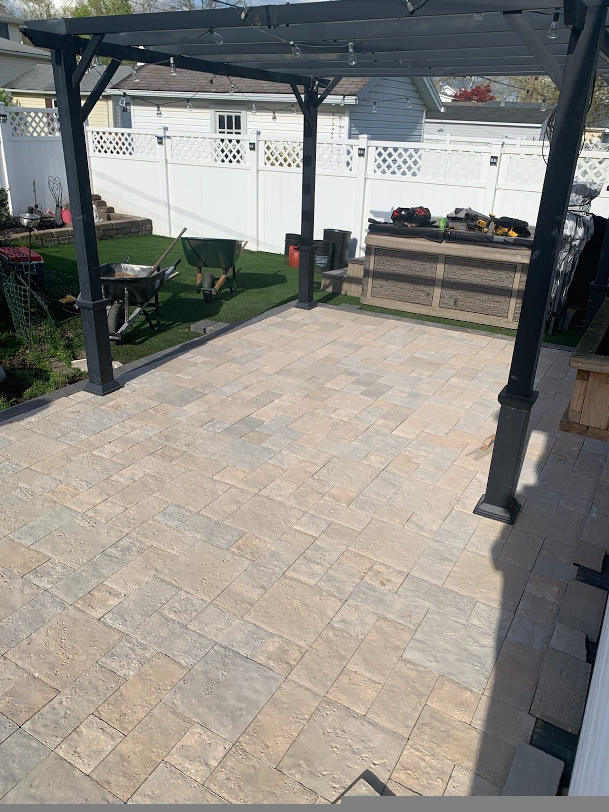 Patio with stone pavers under a pergola, bordered by a white fence and raised planter boxes.