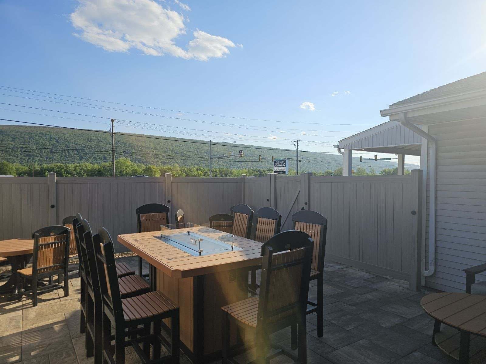 Patio with wooden dining table and chairs beside a house, overlooking a fenced yard and hills under a blue sky