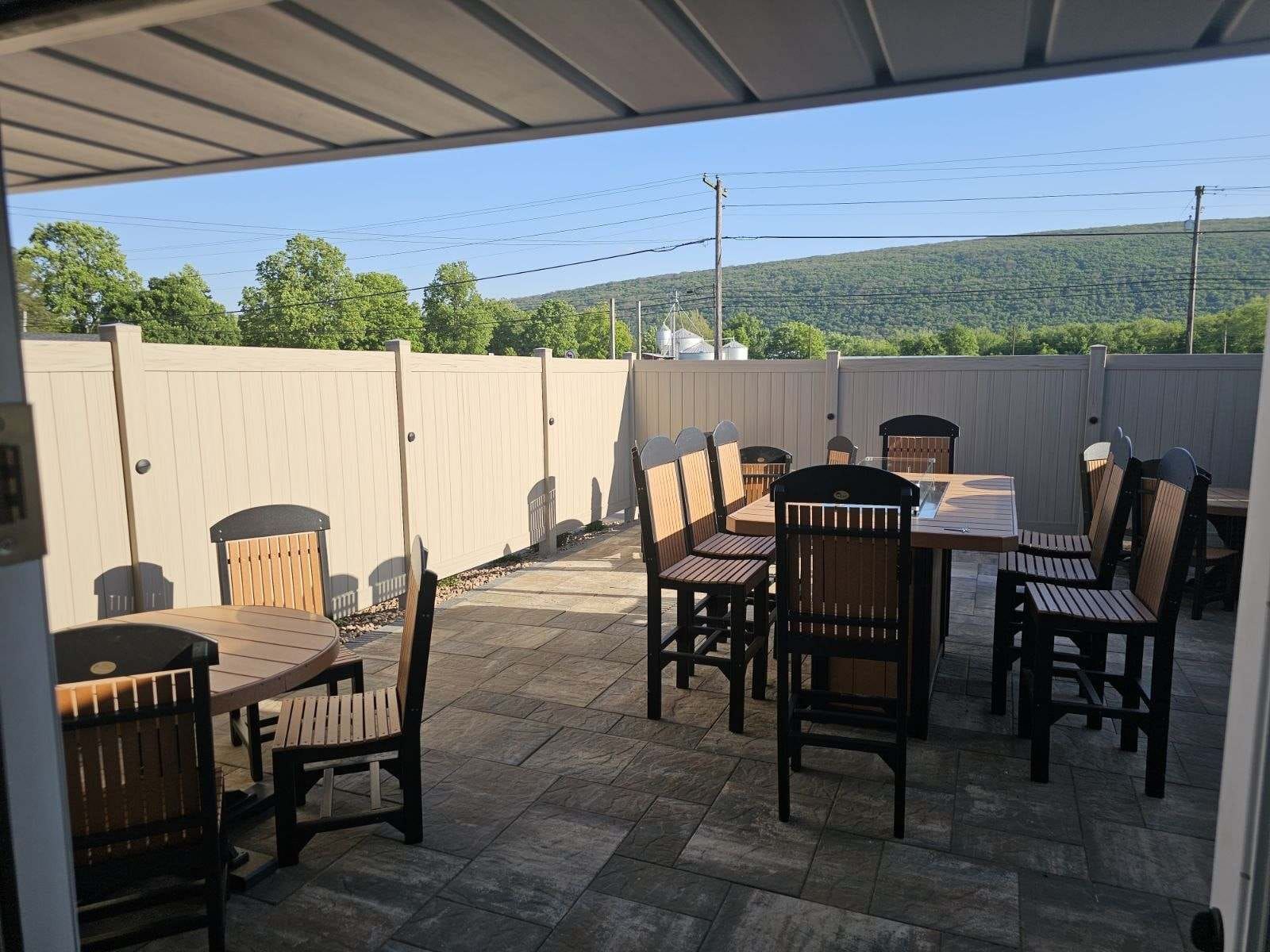 Covered patio with wooden dining tables and chairs on a stone-paver patio, bordered by a white fence.