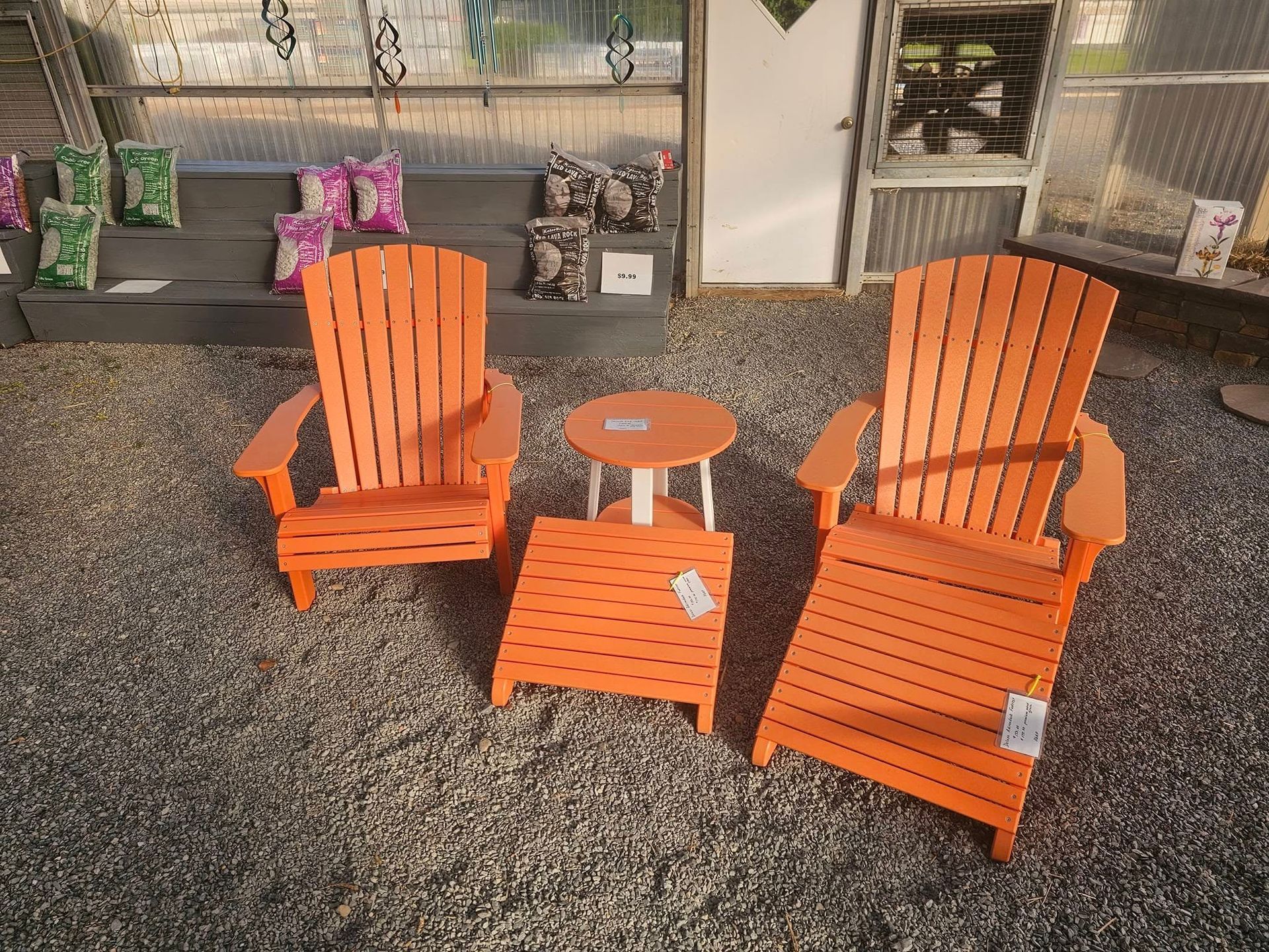 Two orange Adirondack chairs and a small round table on a patio with potted plants and a screen wall