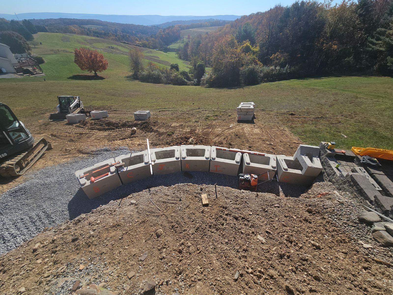 Curved cinder block retaining wall under construction on a gravel site with rolling hills in the background
