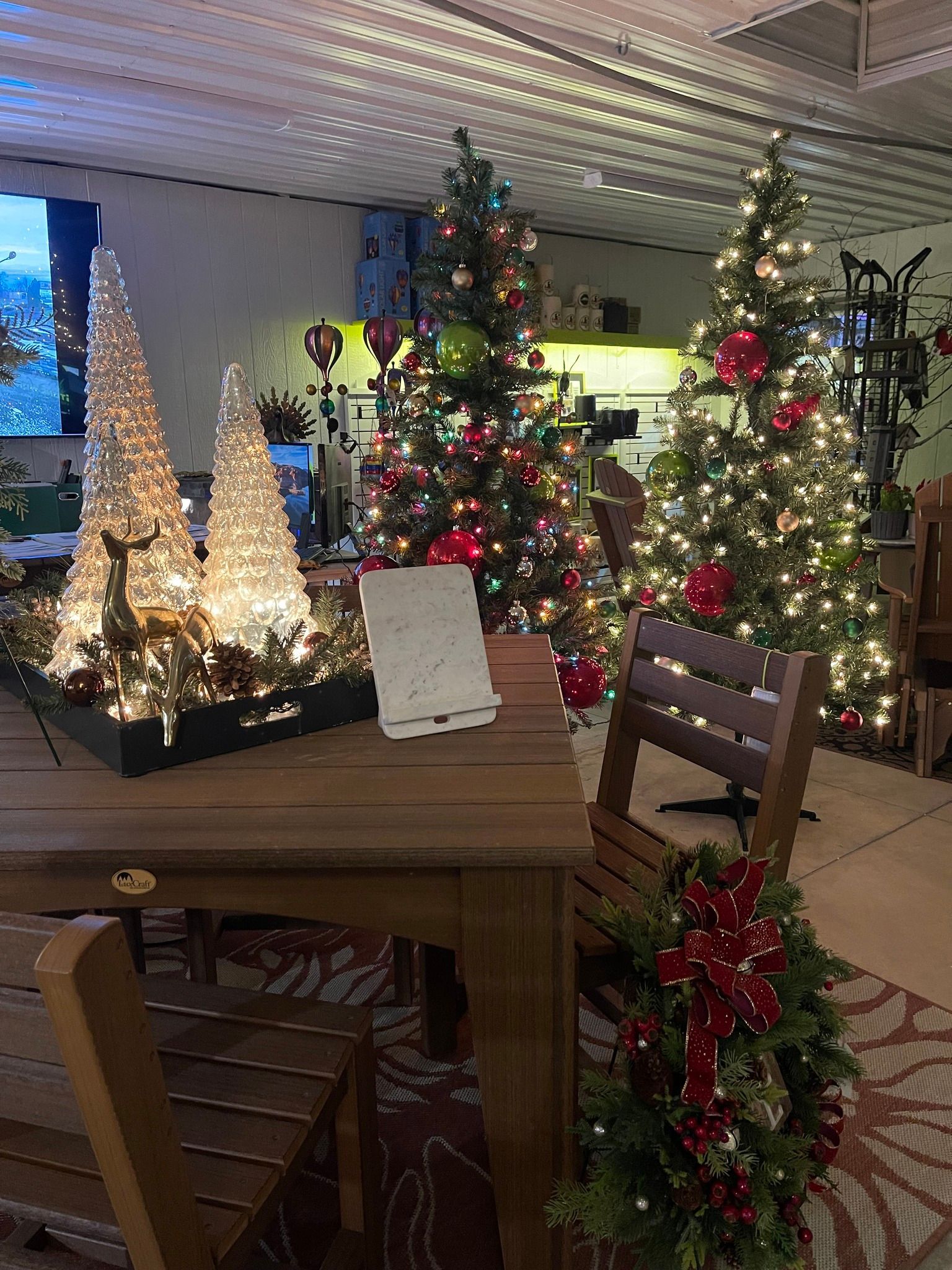 Indoor holiday display with decorated Christmas trees, lights, and a wreath-covered chair by a wooden table.
