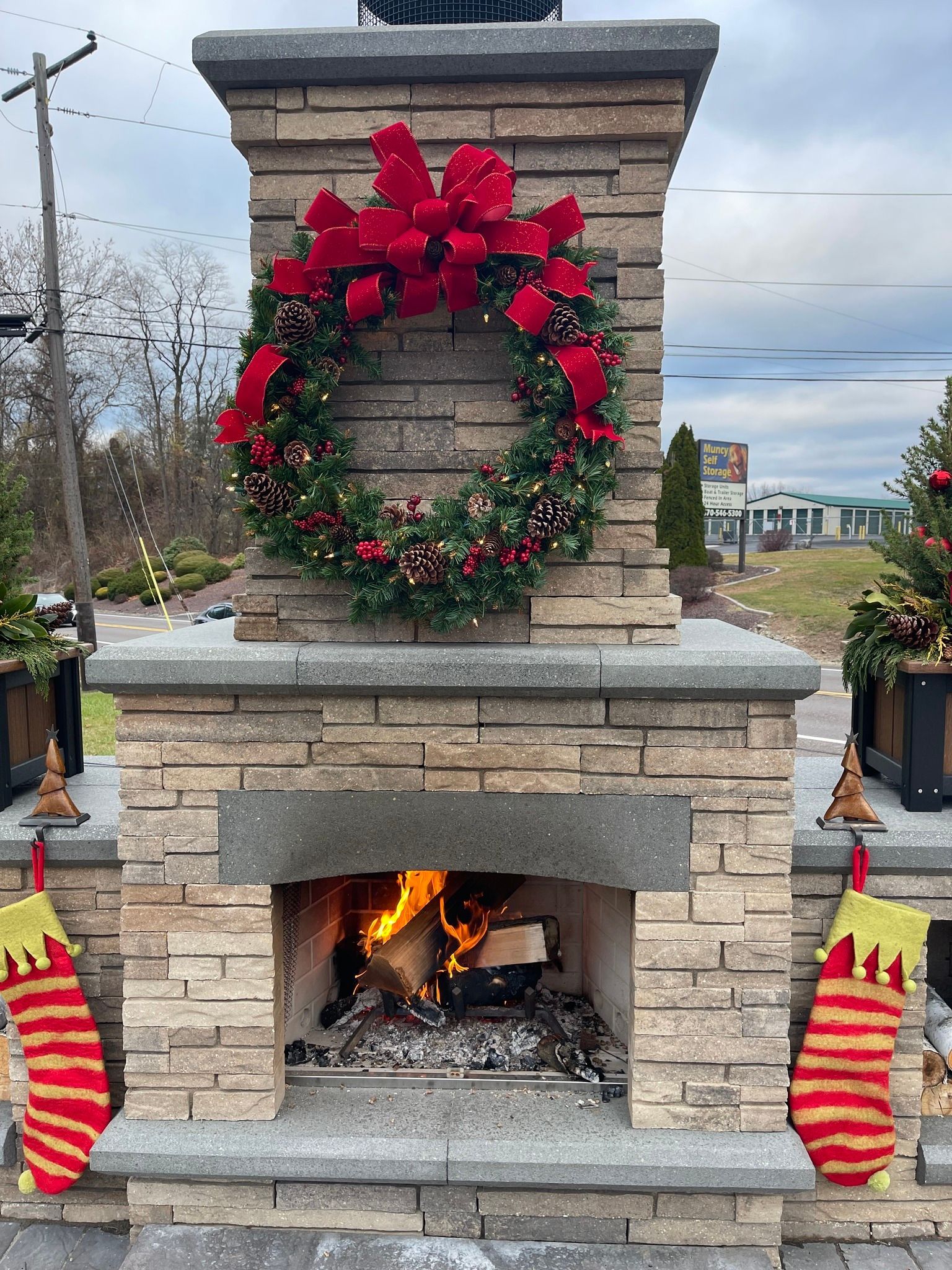 Outdoor stone fireplace decorated with a red bow and wreath, with stockings hanging beside a lit fire.