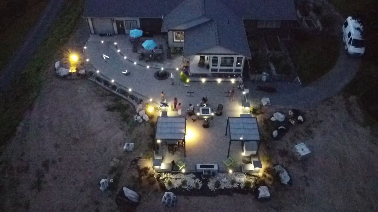 Aerial view of a lit outdoor gathering at night with tables, chairs, and people around a central building.