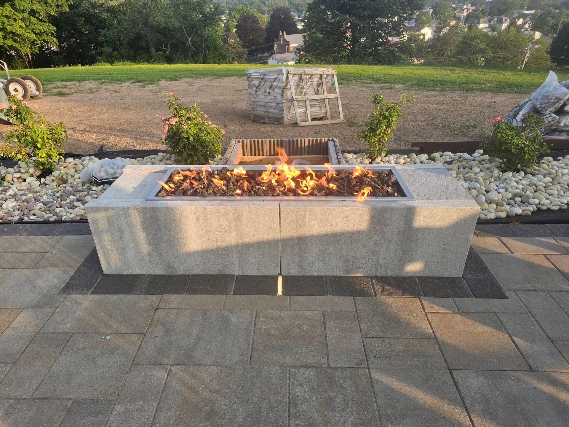 Outdoor fire pit with flames in a stone patio, surrounded by rocks and greenery at dusk