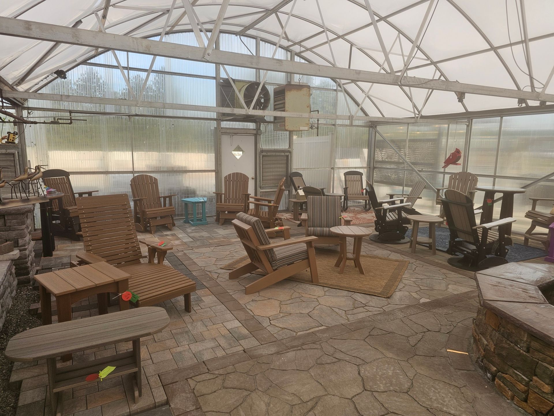 Old greenhouse-like room filled with rustic wooden chairs, tables, and stacked furniture beneath a glass roof