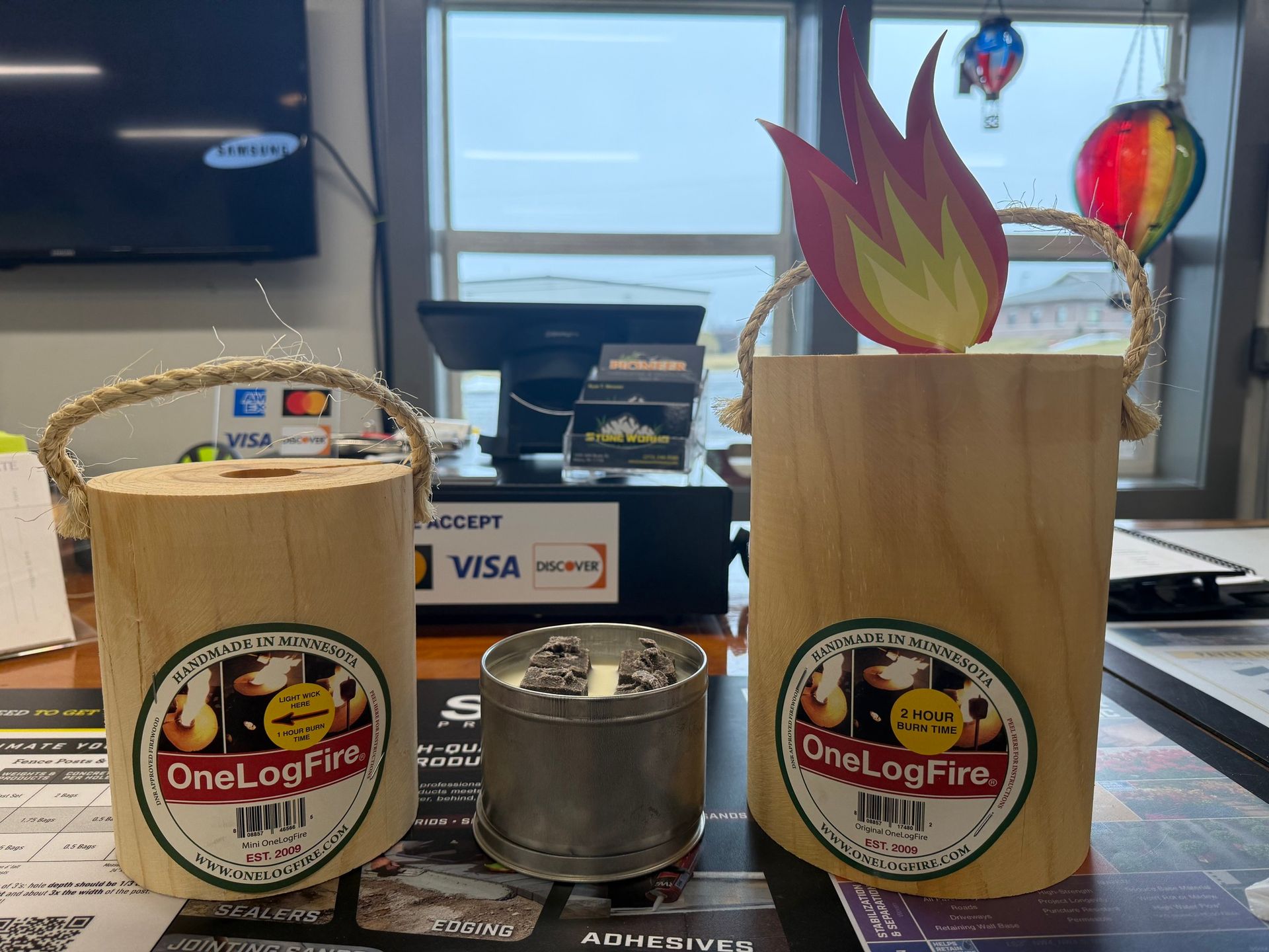 Two OneLogFire tins, a small metal stove, and flame-shaped cutouts on a tabletop near a window.