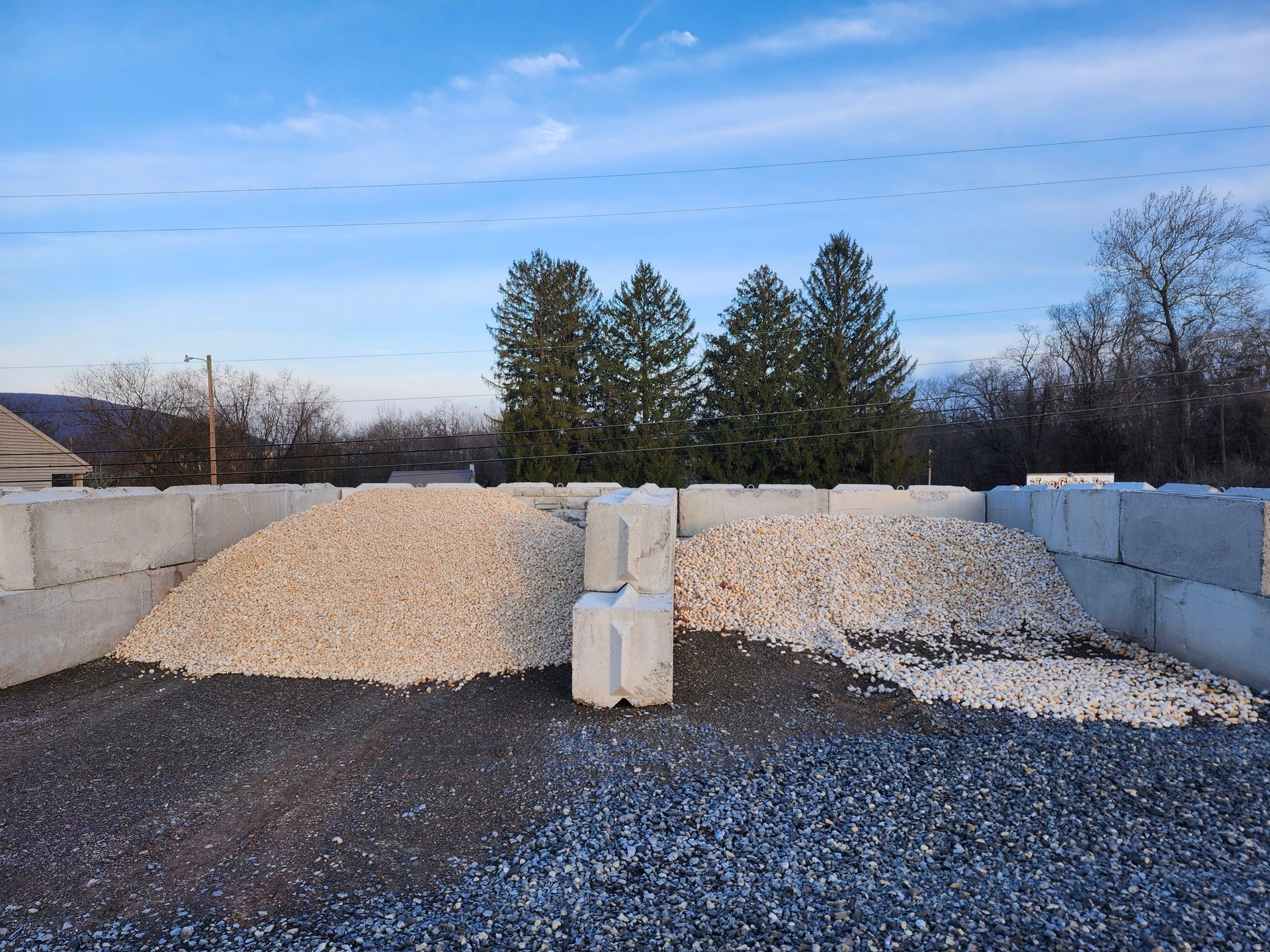 Gravel and white stone piles in a concrete storage yard under a blue sky, with trees in the background