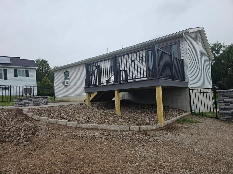 Raised deck with black railings and yellow supports beside a gravel driveway and house.