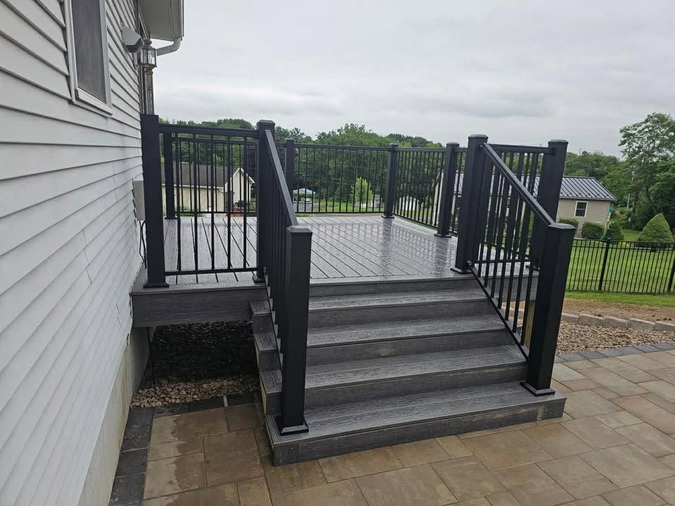 Black deck stairs with railings beside a house, leading to a raised patio over a paved area.