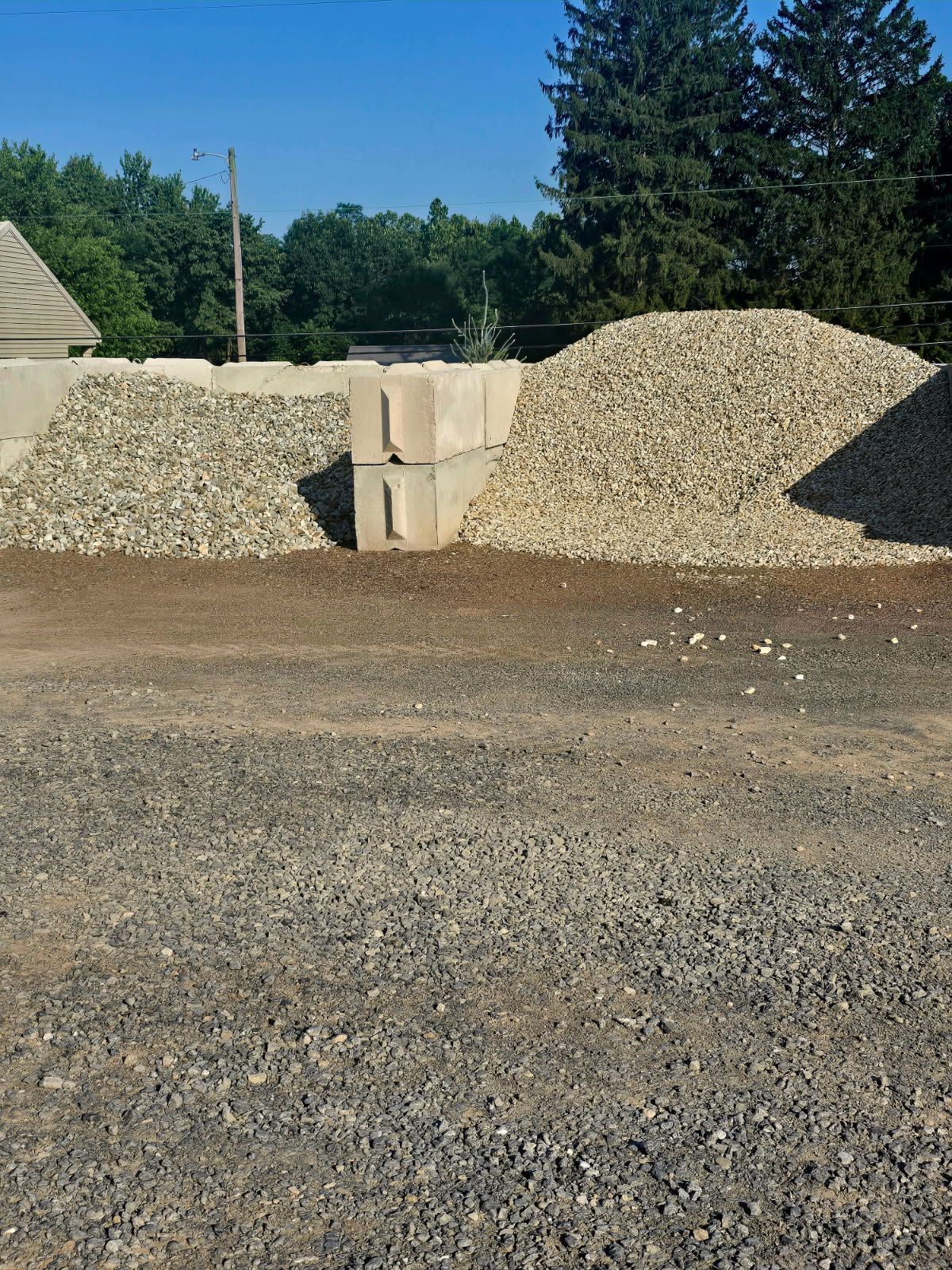 Gravel piles and concrete blocks in a yard under a blue sky, with trees in the background