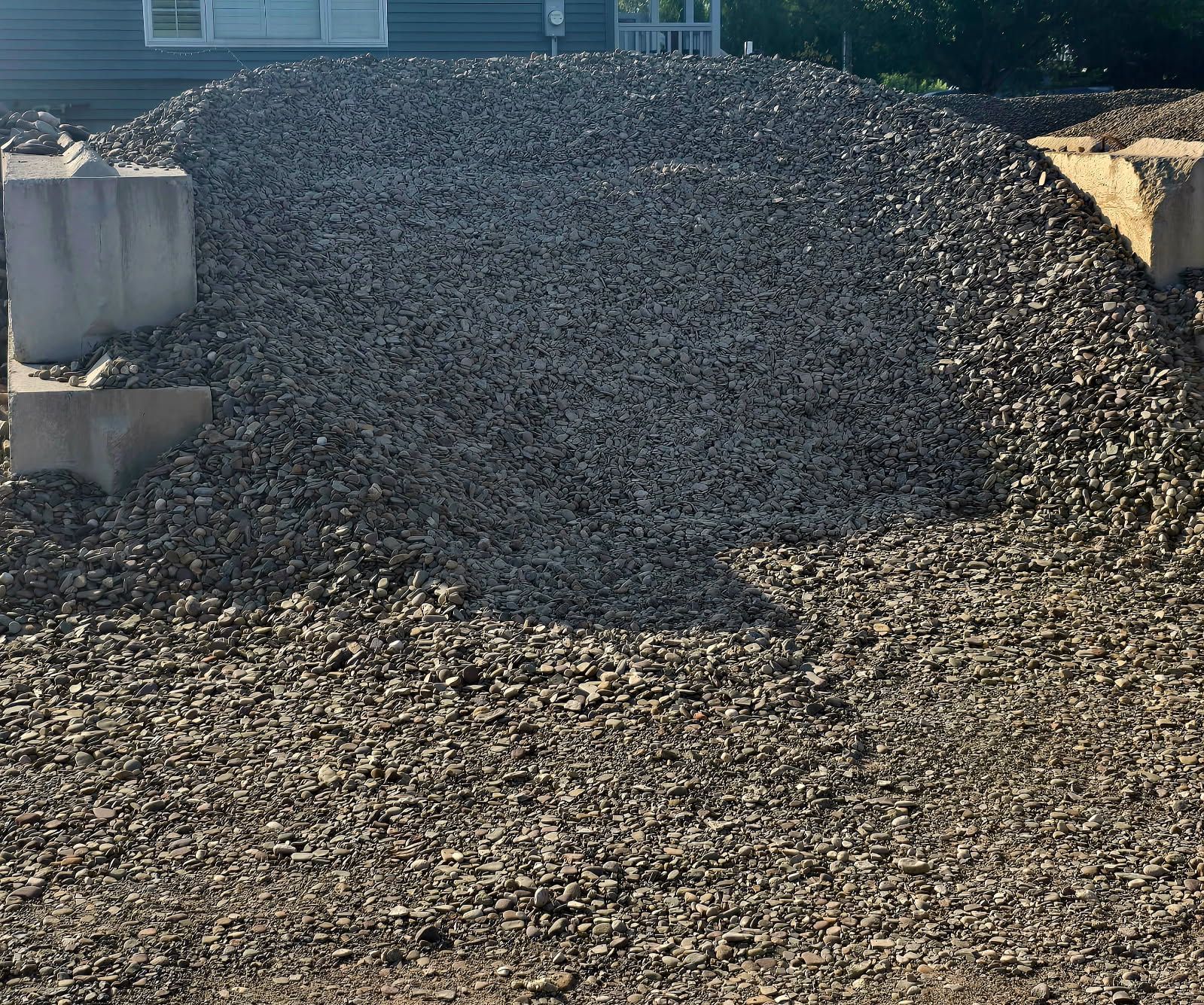 A gravel-covered construction site with a sloped mound between concrete retaining walls and blue containers.