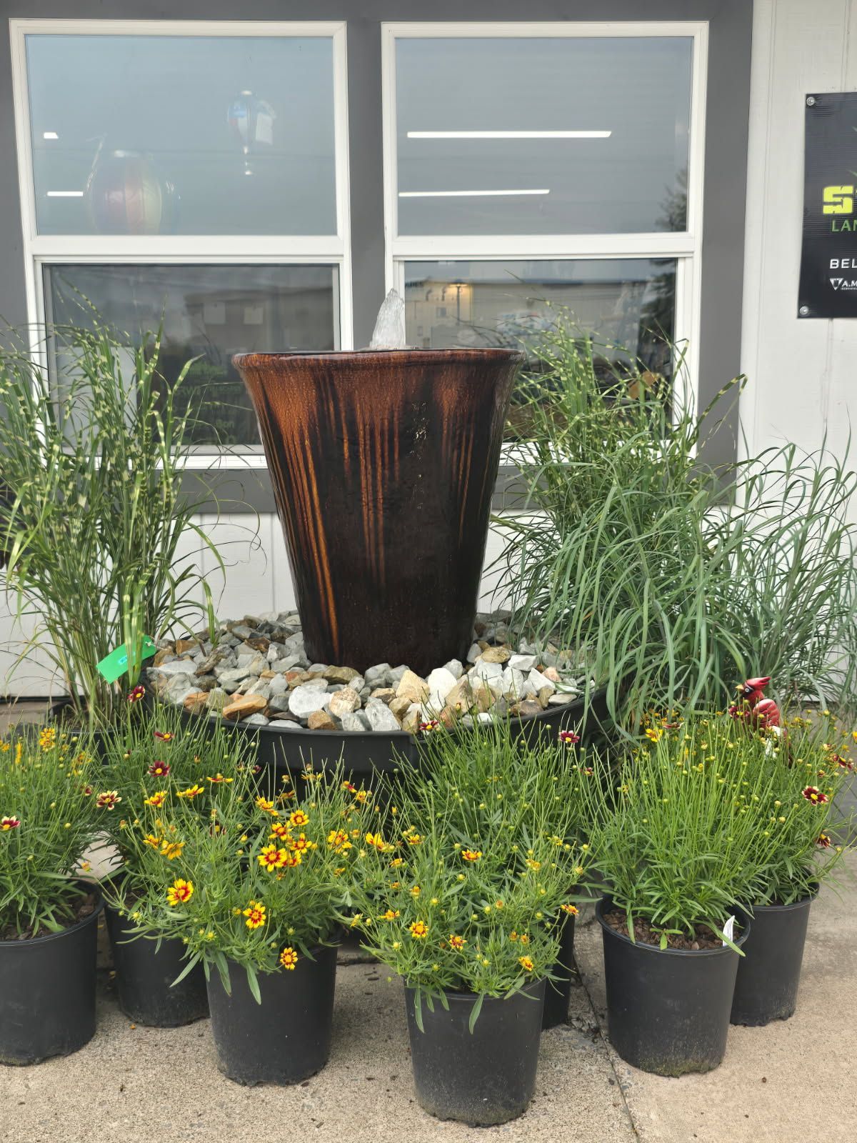 Decorative metal fountain surrounded by potted flowers and grasses in front of a building window