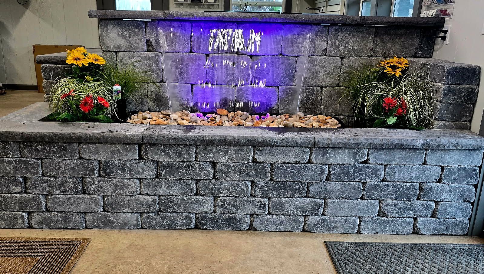Stone garden fountain with purple-lit water, flowers, and a low stacked-stone wall